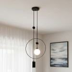 Lampa wisząca Trosti 2-punktowa w kolorze czarnym, z okrągłym ramieniem oraz przezroczystą kulą świetlną. Zawieszona na dwóch cienkich linkach, z dekoracyjnymi kulkami. Tło jasne, z zasłonami i obrazem w stonowanej kolorystyce.