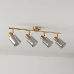 Lampa sufitowa Santia z czterema reflektorami, wykonana z mosiądzu i dymionego szkła. Reflektory mają cylindryczny kształt, a całość zamontowana jest na złotej listwie. Produkt wisi pod sufitem na jednolitym tle.
