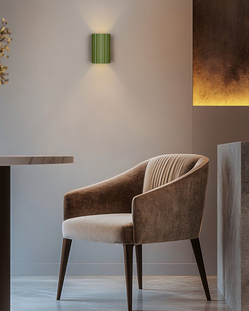 Kinkiet Imaria 1xG9 w kolorze zielonym, o cylindrycznym kształcie z pionowymi żłobieniami, zamontowany na ścianie. W tle widoczny elegancki fotel w odcieniach brązu oraz minimalistyczne wnętrze.