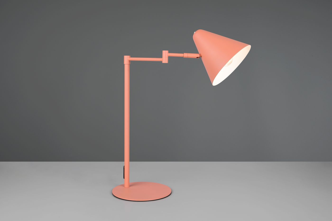 Lampa stołowa Cosima w kolorze morelowym z metalowym stelażem. Klosz w kształcie stożka, zamocowany na regulowanym ramieniu. Ustawiona na okrągłej podstawie, w perspektywie frontalnej. Tło szare.