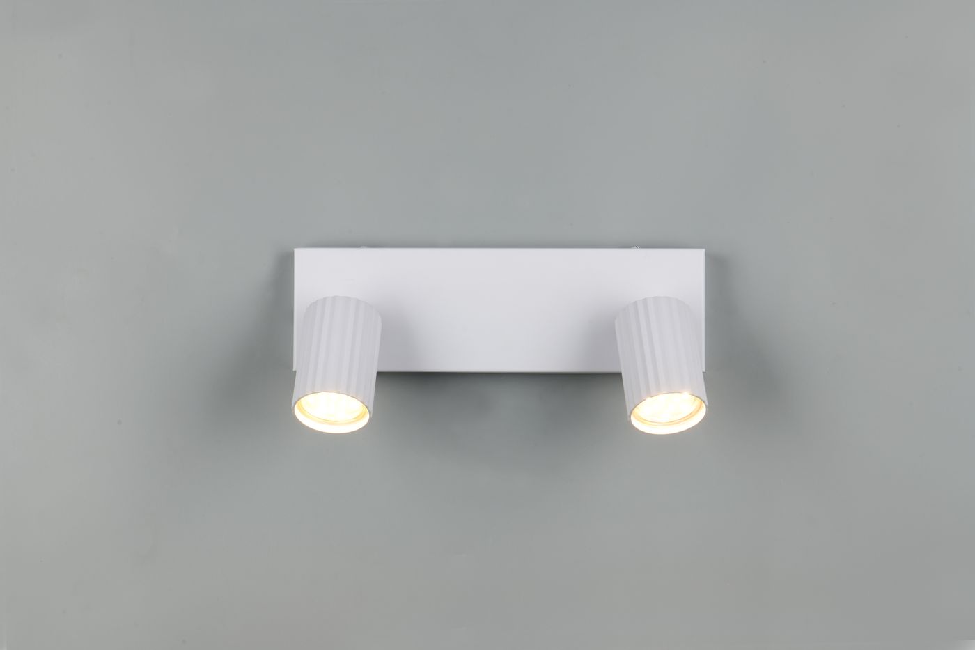 Lampa sufitowa Soka w kolorze białym z dwoma reflektorami na wysięgnikach. Reflektory mają teksturowane boki i emitują ciepłe światło. Produkt zamontowany na prostokątnej podstawie, widok frontalny.