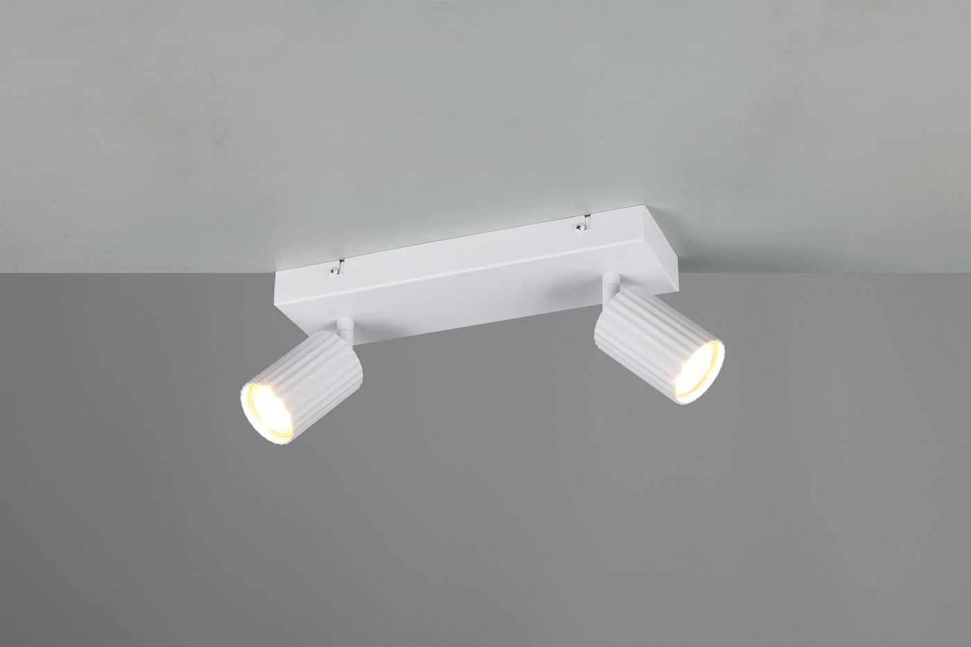 Lampa sufitowa Soka z dwoma reflektorami GU10 w białym kolorze, zamontowana na prostokątnej podstawie. Reflektory mają teksturowaną powierzchnię i są ustawione pod różnymi kątami. Tło w odcieniu szaro-beżowym.