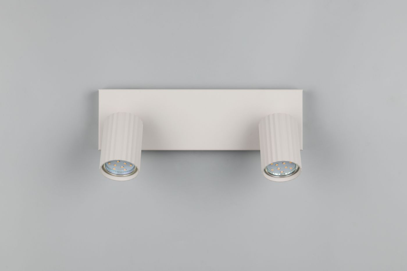Lampa sufitowa Soka w kolorze szarym, z dwoma reflektorami GU10. Kształt cylindryczny z delikatnymi żłobieniami. Montaż na płaskiej podstawie, widoczne źródła światła. Tło w stonowanej szarości.