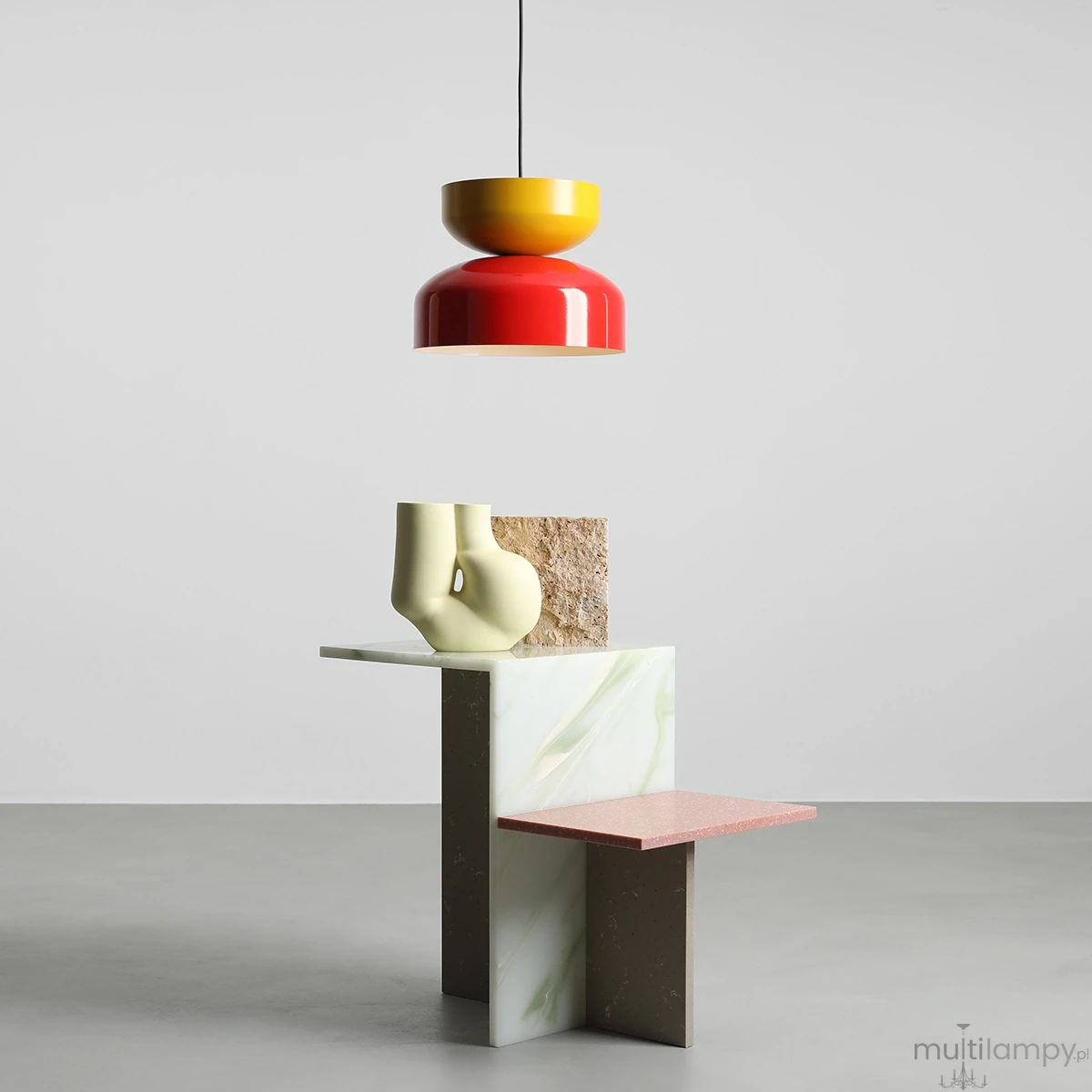 Lampa wisząca Tula w kolorach czerwonym i musztardowym, z kloszem w kształcie stożka. Zawieszona na cienkim przewodzie, umieszczona nad dekoracyjnym stolikiem z geometrycznymi elementami. Tło jasne, minimalistyczne.
