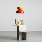 Lampa wisząca Tula w kolorach czerwonym i musztardowym, z kloszem w kształcie stożka. Zawieszona na cienkim przewodzie, umieszczona nad dekoracyjnym stolikiem z geometrycznymi elementami. Tło jasne, minimalistyczne.