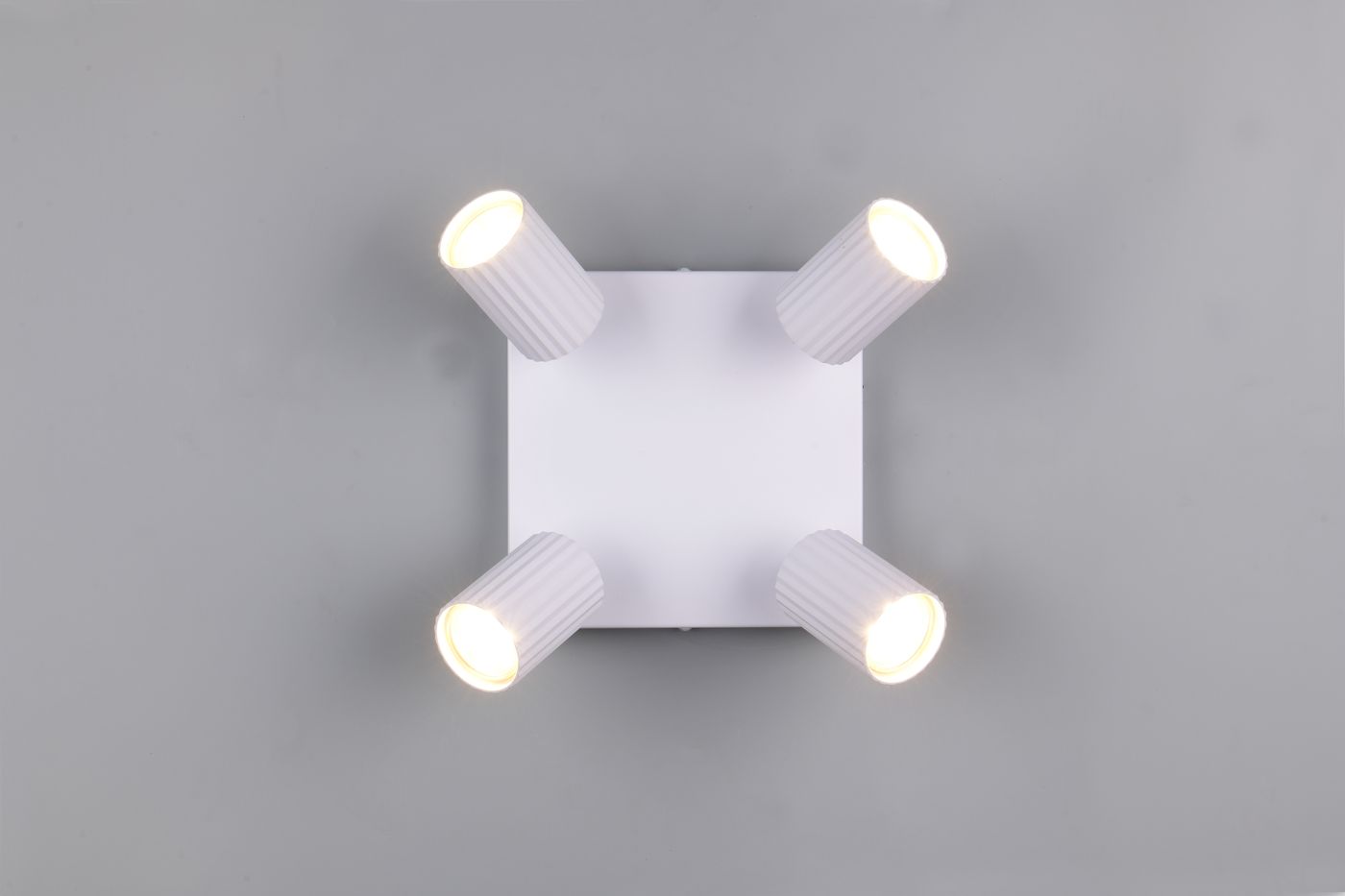 Lampa sufitowa Soka w kolorze białym z czterema regulowanymi reflektorami GU10. Ujęcie z góry, produkt zamontowany na płaskiej powierzchni. Reflektory mają rowkowaną fakturę, emitują ciepłe światło. Tło szare, proste.