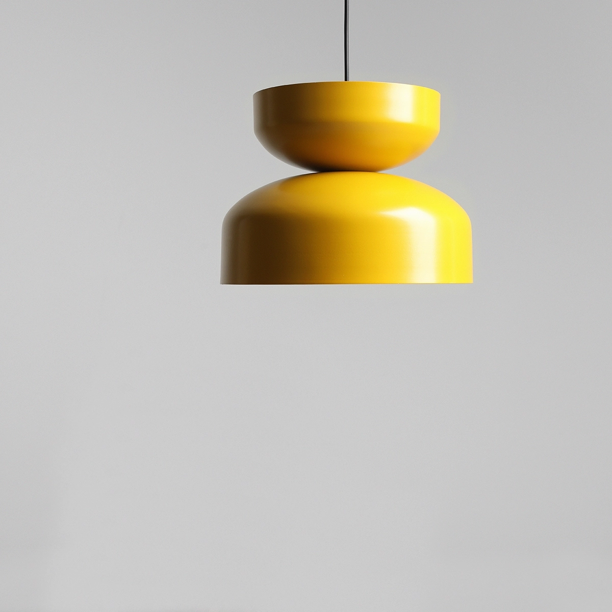 Lampa wisząca Tula 1xE27 w kolorze musztardowym, o nowoczesnym kształcie składającym się z dwóch połączonych elementów. Zawieszona na czarnym przewodzie, na jednolitym, jasnym tle.