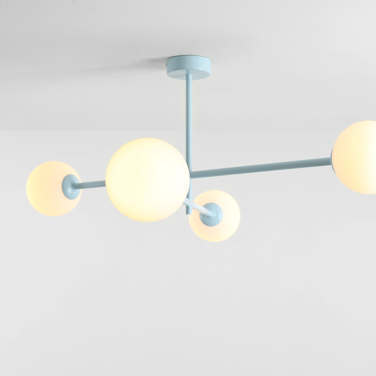 Lampa sufitowa Pinne w kolorze błękitnym, z czterema okrągłymi kloszami w kolorze mlecznym, zawieszona na cienkich metalowych ramionach. Ujęcie z góry pokazujące konstrukcję i detale mocowania sufitowego.