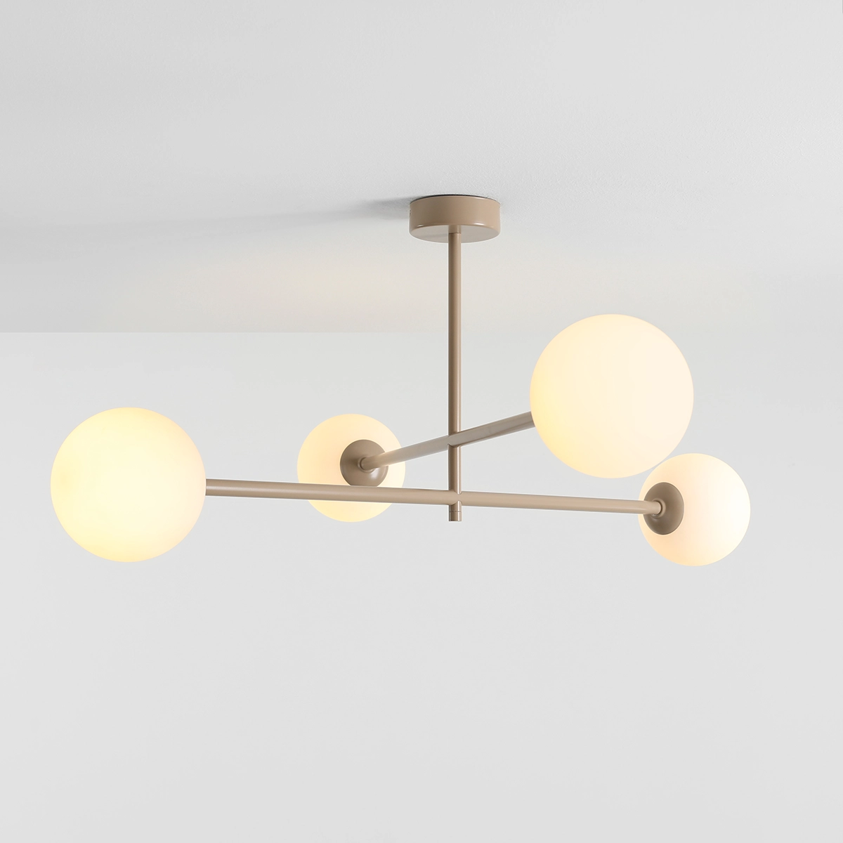 Beżowa lampa sufitowa Pinne z czterema kulistymi kloszami, wykonana z metalu, z zamontowanym centralnym mocowaniem. Klosze w kolorze mlecznym, emitujące miękkie światło, umieszczone na asymetrycznych ramionach. Ujęcie z dołu.