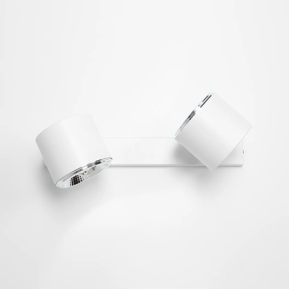 Kinkiet 2-punktowy w kolorze białym, zamontowany na prostokątnej podstawie. Dwa okrągłe klosze z metalicznymi detalami, skierowane w różnych kierunkach. Prosty, nowoczesny design na jednolitym tle.