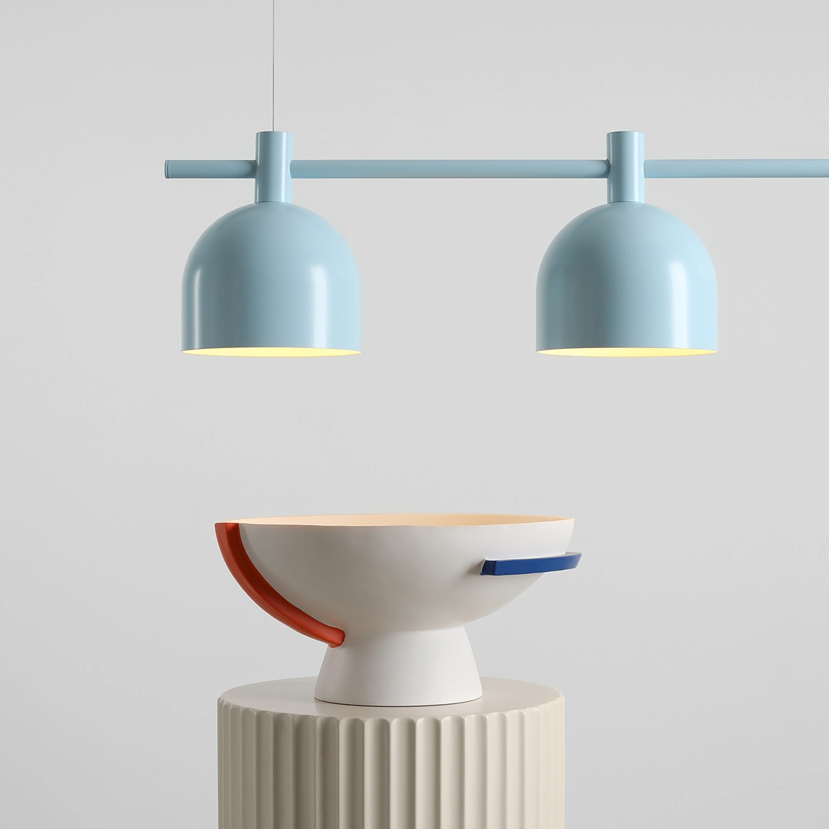 Lampa wisząca Beryl z trzema kloszami E27 w błękitnym kolorze, zawieszona na poziomej rurze. W tle widoczna dekoracyjna misa na okrągłej podstawie. Tło jasnoszare, minimalistyczne.