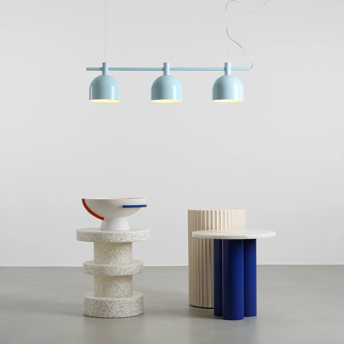 Lampa wisząca Beryl w kolorze niebieskim z trzema kloszami E27, zawieszona na prostym stelażu. Produkt wisi nad minimalistycznym wnętrzem z nowoczesnymi meblami, w tym okrągłym stołem z niebieskimi nogami i dekoracyjną misą.