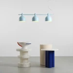 Lampa wisząca Beryl w kolorze niebieskim z trzema kloszami E27, zawieszona na prostym stelażu. Produkt wisi nad minimalistycznym wnętrzem z nowoczesnymi meblami, w tym okrągłym stołem z niebieskimi nogami i dekoracyjną misą.