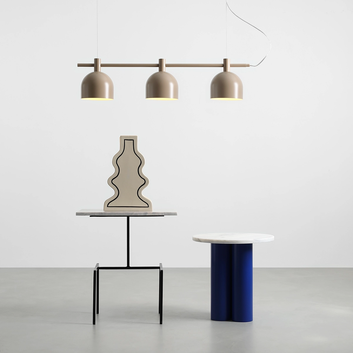 Lampa wisząca Beryl w beżowym kolorze z trzema kloszami, zawieszona na metalowej rurze. W tle widoczna dekoracyjna figura na stoliku z marmurowym blatem oraz okrągły stół w niebieskim kolorze. Tło jasne, minimalistyczne.