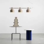 Lampa wisząca Beryl w beżowym kolorze z trzema kloszami, zawieszona na metalowej rurze. W tle widoczna dekoracyjna figura na stoliku z marmurowym blatem oraz okrągły stół w niebieskim kolorze. Tło jasne, minimalistyczne.