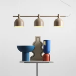 Beżowa lampa wisząca Beryl z trzema kloszami E27, zawieszona nad stołem. W tle kolorowe dekoracje: niebieska misa, ceramiczne figury w odcieniach czerwieni i bieli. Proste, jasne tło podkreśla nowoczesny design lampy.