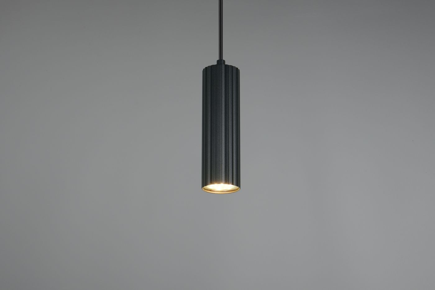 Lampa wisząca Soka w kolorze czarnym z pojedynczym źródłem światła GU10. Cylindryczny kształt z fakturowaną powierzchnią, zawieszona na cienkim kablu, emituje światło z dolnej części. Tło jasnoszare, minimalistyczne.