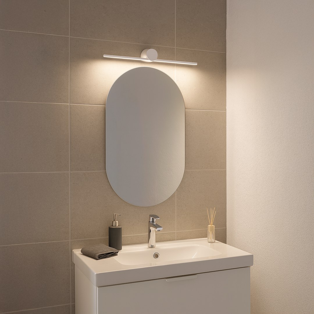 Biały kinkiet Artist L40,5 LED 6W 4000K IP44, zamontowany nad owalnym lustrem. Oświetlenie skierowane na lustro, otoczenie w neutralnych odcieniach szarości. Kinkiet ma minimalistyczny design, zamocowany na gładkiej, jasnej ścianie.