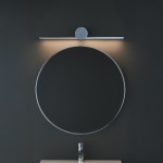 Kinkiet Artist L40,5 LED 6W 4000K IP44 w kolorze chromu, zamontowany nad okrągłym lustrem. Oświetlenie skierowane w dół, na tle ciemnej ściany. Stylowy, minimalistyczny design z okrągłym elementem centralnym.