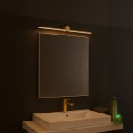 Kinkiet Artist L60,5 LED 9W 4000K IP44 w kolorze złotym, zamontowany nad lustrem w łazience. Posiada prostą, liniową formę, oświetlającą przestrzeń. Widoczna elegancka bateria umywalkowa i zielona butelka na blacie. Tło w ciemnym odcieniu.