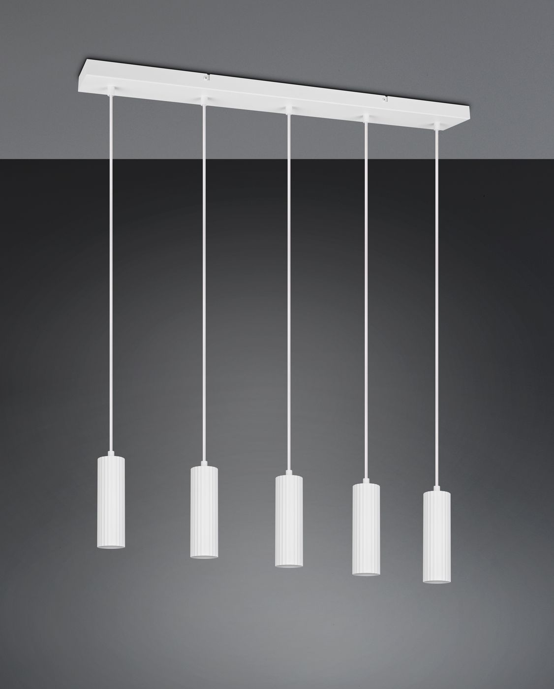 Lampa wisząca Soka z pięcioma oprawami GU10, w białym kolorze. Cylindryczne klosze z rowkowaną fakturą zawieszone na cienkich linkach. Montowana na prostokątnej podstawie. Tło ciemnoszare, podkreślające elegancki design.