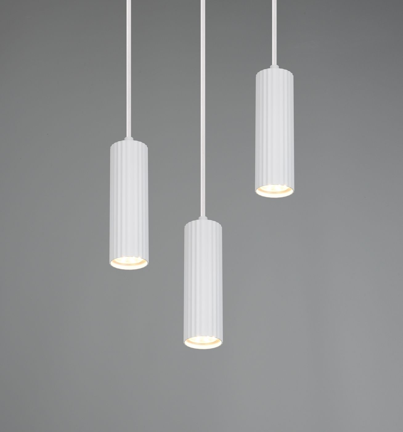 Lampa wisząca Soka z trzema kloszami GU10 w kolorze białym. Klosze mają cylindryczny kształt z pionowymi żłobieniami. Produkt zawieszony na cienkich linkach, na jednolitym szarym tle.