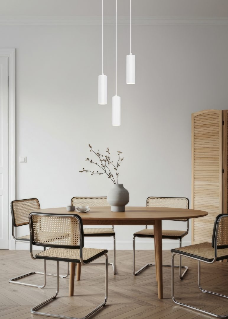 Lampa wisząca Soka w kolorze białym, z trzema cylindrycznymi kloszami, zawieszona na cienkich linkach. Klosze mają żłobioną fakturę. W tle drewniany stół z ceramiczną wazonem i krzesłami z plecionkami. Ściana w neutralnym odcieniu.