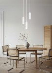 Lampa wisząca Soka w kolorze białym, z trzema cylindrycznymi kloszami, zawieszona na cienkich linkach. Klosze mają żłobioną fakturę. W tle drewniany stół z ceramiczną wazonem i krzesłami z plecionkami. Ściana w neutralnym odcieniu.