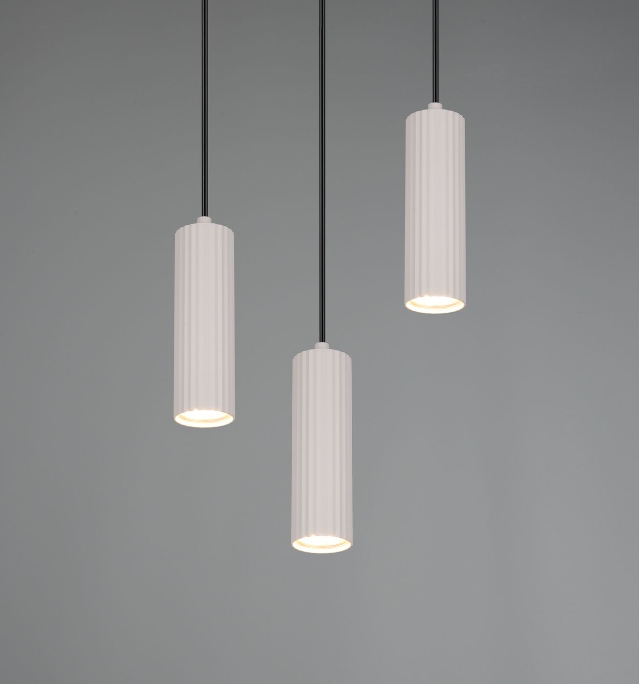 Lampa wisząca Soka z trzema cylindrycznymi kloszami w kolorze szarym, zawieszonymi na czarnych linkach. Klosze mają żłobioną fakturę i emitują ciepłe światło. Produkt zawiera oprawy na żarówki GU10. Tło w odcieniach szarości.