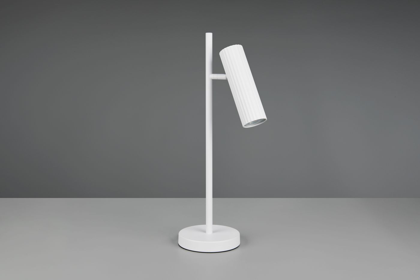 Lampa stołowa Soka w kolorze białym z oprawą 1xGU10, z okrągłą podstawą i prostym, pionowym stelażem. Kluczowy element to cylindryczny klosz z rowkowaną powierzchnią, nachylony pod kątem. Produkt na szarym tle.