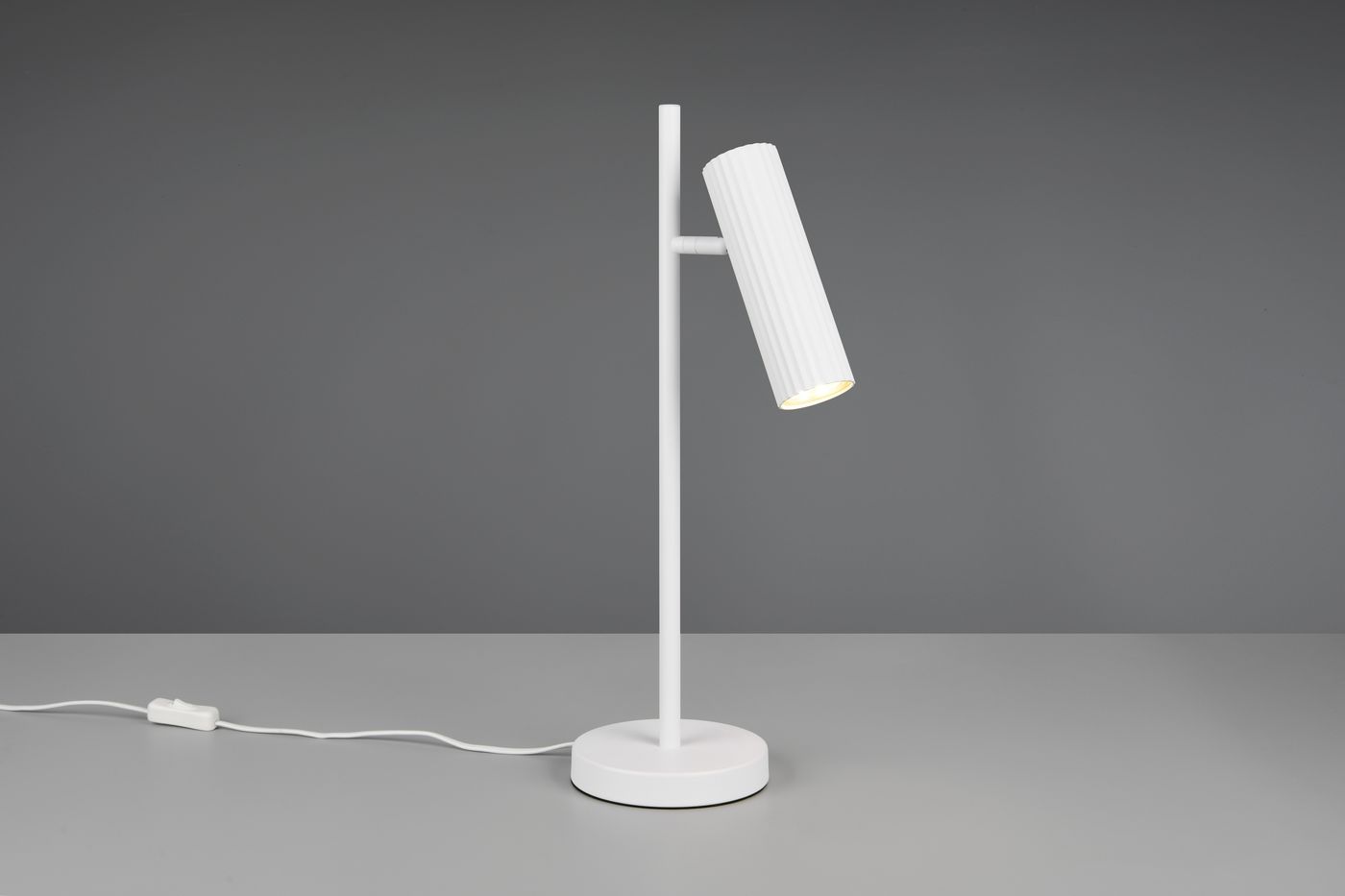 Lampa stołowa Soka w kolorze białym, z okrągłą podstawą i pionowym stelażem. Główka lampy w formie cylindrycznej, żłobkowanej, skierowana lekko w dół. Widoczny przewód z przełącznikiem. Ujęcie z boku na jednolitym tle.