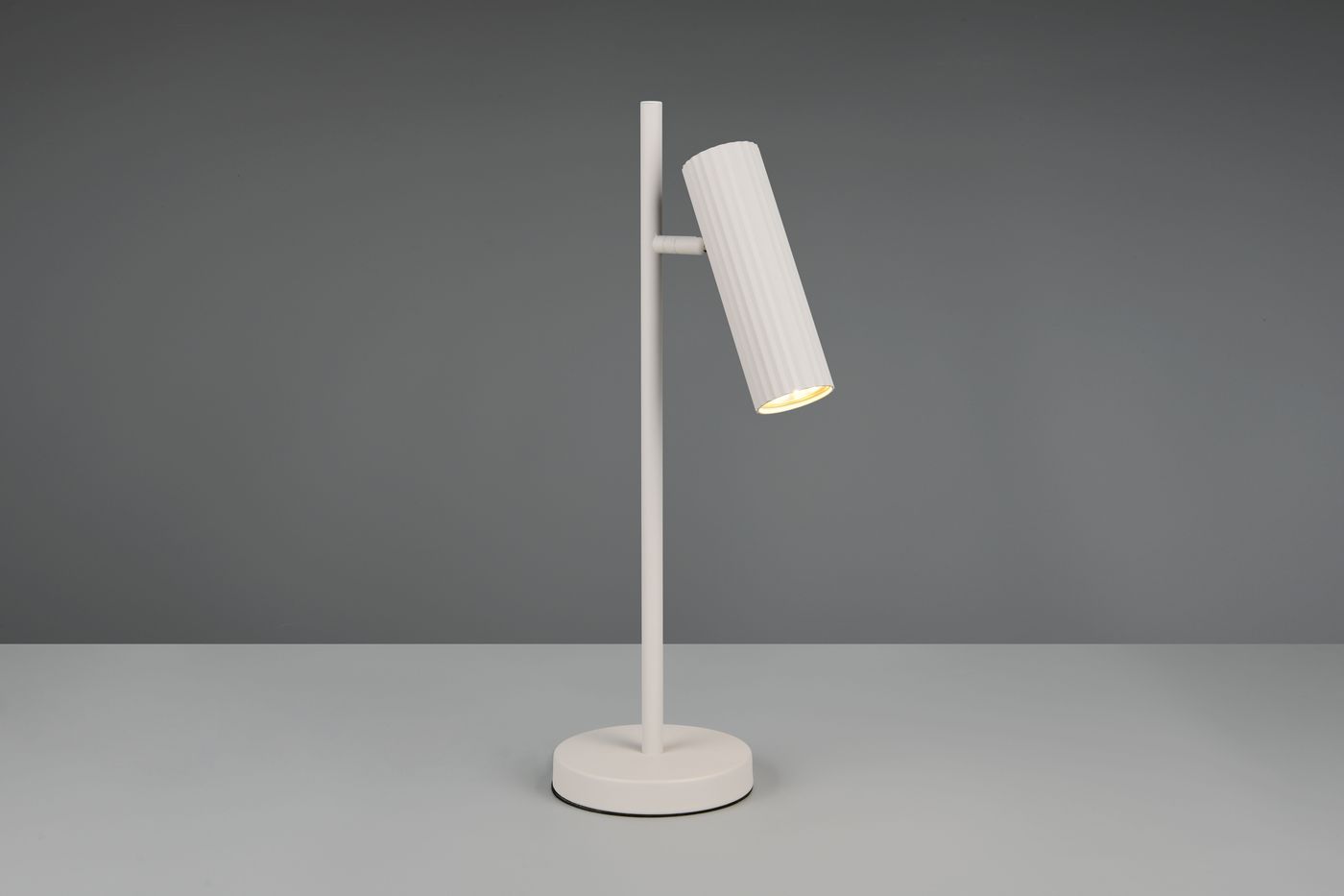 Lampa stołowa Soka w kolorze szarym z podstawą okrągłą. Stylizowany klosz w formie walca z teksturą, zamontowany na prostym stelażu. Ujęcie frontalne, lampa ustawiona w pozycji stojącej, skierowana lekko w dół.