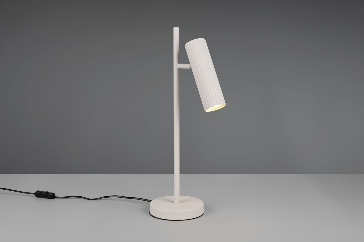 Lampa stołowa Soka w kolorze szarym z regulowanym kloszem w formie cylindrycznej, umieszczona na okrągłej podstawie. Ujęcie z boku, z widocznym kablem z włącznikiem. Kluczowa funkcja to możliwość skierowania światła.
