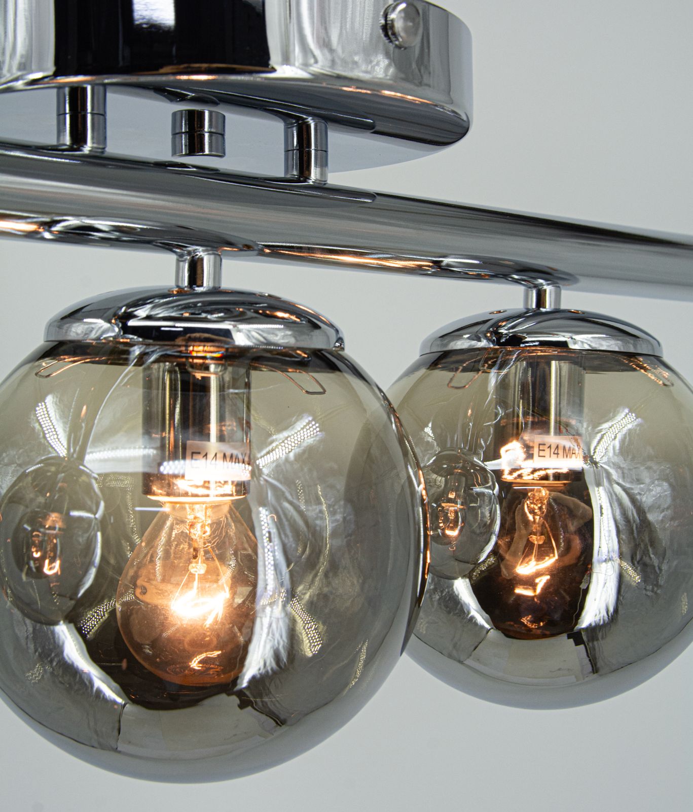 Lampa sufitowa Argo z trzema oprawami E14 w chromowanym wykończeniu. Okrągłe, przezroczyste klosze w kolorze szarym, zawieszone na metalowej ramie. Widoczne żarówki emitujące ciepłe światło. Produkt zamontowany na suficie.