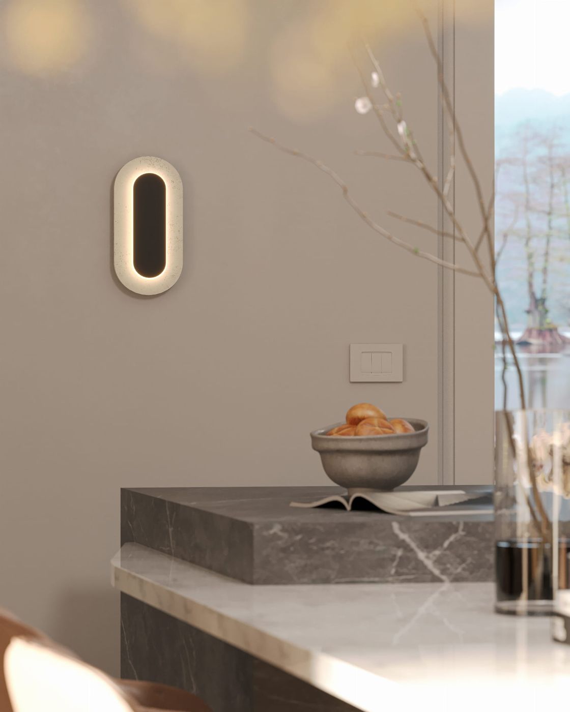 Kinkiet Travertino Oval z naturalnego kamienia, w kształcie owalnym, z wbudowanym źródłem LED 10W. Zainstalowany na ścianie, emituje ciepłe światło, w otoczeniu minimalistycznego wnętrza z jasnymi tonami. W tle widoczny stół z kamiennym blatem.