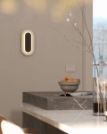 Kinkiet Travertino Oval z naturalnego kamienia, w kształcie owalnym, z wbudowanym źródłem LED 10W. Zainstalowany na ścianie, emituje ciepłe światło, w otoczeniu minimalistycznego wnętrza z jasnymi tonami. W tle widoczny stół z kamiennym blatem.