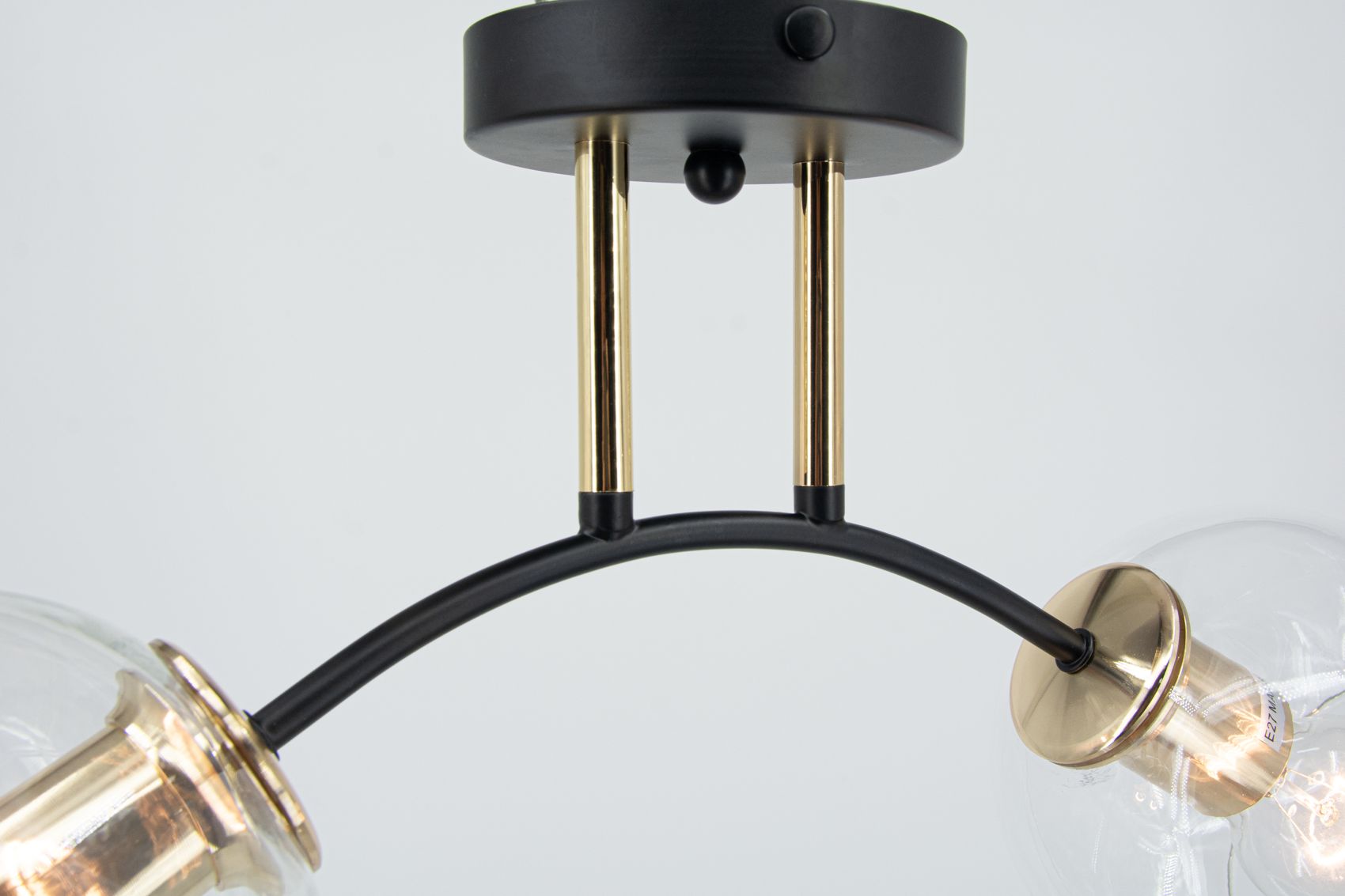 Lampa sufitowa Elba z dwoma oprawami E27, w kolorze czarnym i złotym. Ujęcie z boku, pokazujące okrągły sufitowy uchwyt oraz łukowate rury. Widać przezroczyste klosze, które eksponują żarówki. Tło białe, proste.