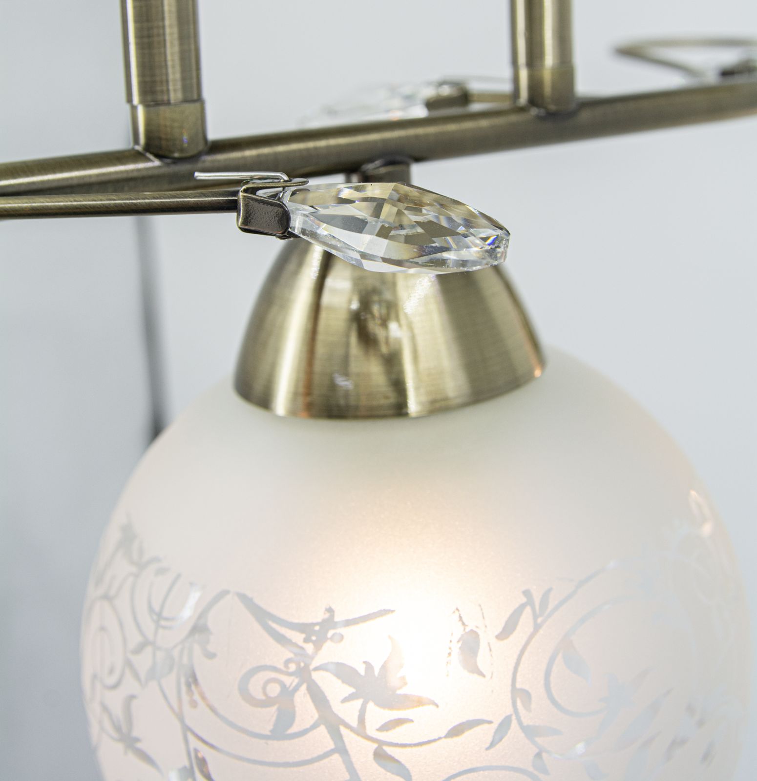 Lampa sufitowa Olivier w kolorze patyny, z trzema oprawami E27. Posiada szklane klosze z matowym wykończeniem i dekoracyjnym wzorem. Zbliżenie na detale mocowania i kryształowe elementy ozdobne.