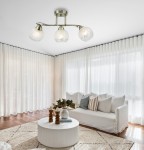 Lampa sufitowa Olivier w kolorze patynowym z trzema kloszami z matowego szkła, umieszczona na metalowej konstrukcji. Klosze mają delikatny wzór i emitują ciepłe światło, zawieszona w jasnym wnętrzu z białą sofą i oknami zasłoniętymi firankami.