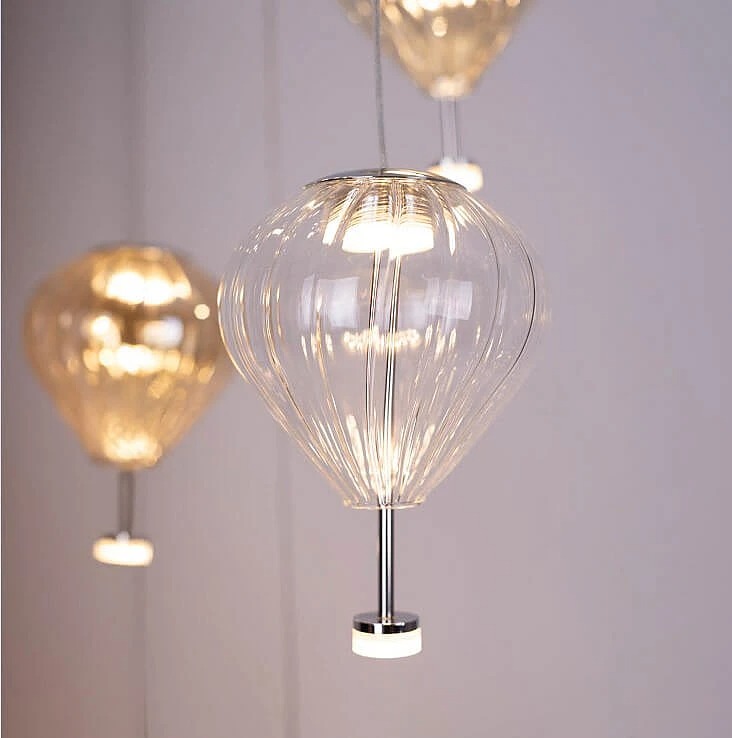 Lampa wisząca Palloncini 2xLED 6W o przezroczystym kloszu w kształcie balonu, z metalowym uchwytem. Zawieszona na cienkim przewodzie, emituje ciepłe światło, idealna do nowoczesnych wnętrz. Tło w jasnym kolorze.