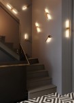 Kinkiet Merano 1xLED 6W w kolorze złotym, zamontowany na ścianie przy schodach. Lampa ma prosty, nowoczesny kształt z źródłem światła, emitującym ciepłe światło. Tło w odcieniach szarości, schody w ciemnym kolorze, geometria podłogi w czarno-białe wzory.