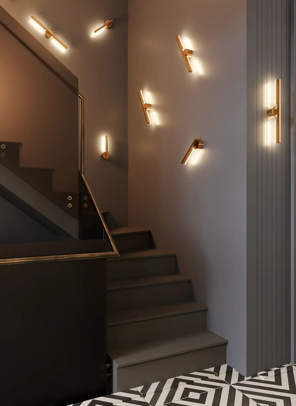Kinkiet Merano 2xLED 10W w kolorze złotym, zamontowany na ścianie schodów. Prosty, cylindryczny kształt, emitujący ciepłe światło. Widoczny na szarym tle, w towarzystwie innych kinkietów, tworzy nowoczesną aranżację oświetleniową.