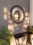 Kinkiet Merano 2xLED 10W w kolorze złotym, umieszczony na ścianie obok okrągłego lustra. Produkt ma minimalistyczny design, z dwiema źródłami światła i metalowymi detalami. W tle widoczna roślinność oraz elegancki stół.