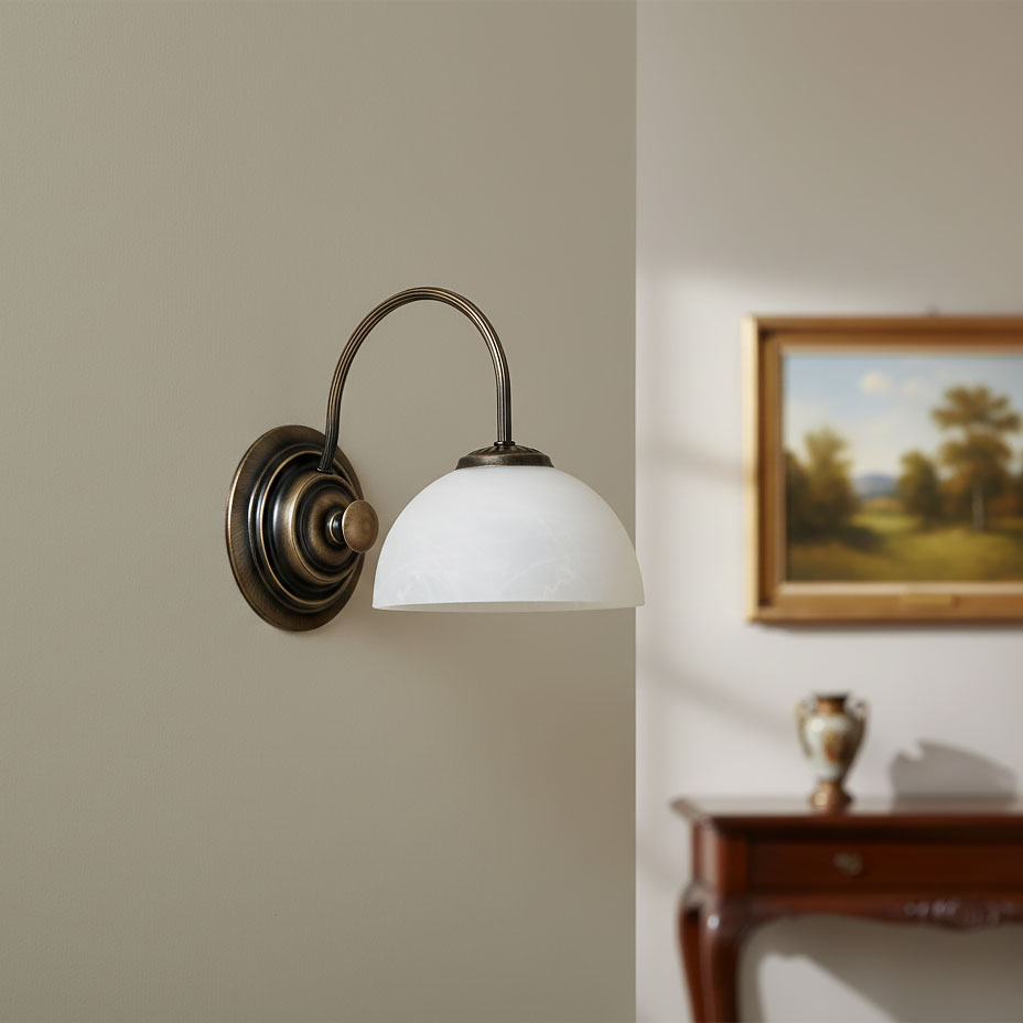 Kinkiet Delos 1xE27 w kolorze patyny, z półkulistym kloszem z mlecznego szkła. Produkt zamontowany na ścianie, z delikatnie wygiętym ramieniem. Tło w neutralnym kolorze, w aranżacji z drewnianym meblem i obrazem w ramie.
