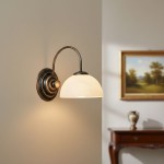 Kinkiet Delos 1xE27 w kolorze patynowym z okrągłą, białą oprawą lampy, umocowany na ścianie. Widok z boku, podkreślający zakrzywioną ramię i detale mocowania. W tle delikatne dekoracje wnętrza.