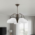 Żyrandol Delos w kolorze patyny z pięcioma kloszami z mlecznego szkła. Ułożenie wiszące, z centralnym mocowaniem, elegancko podkreślający przestrzeń. Tło to jasne wnętrze z zasłonami.