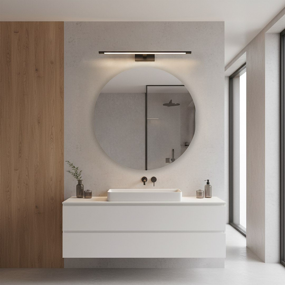 Kinkiet Klimt L60 LED 12W CCT IP44 w kolorze czarnym, zamontowany nad umywalką w nowoczesnej łazience. Oświetlenie emituje przyjemne światło, a jego minimalistyczny design harmonizuje z okrągłym lustrem i jasnym blatem.