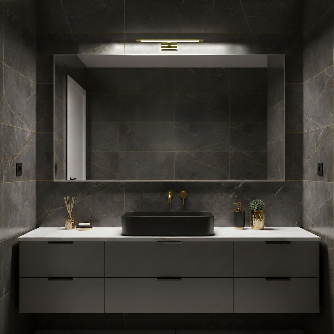 Kinkiet Klimt L40 LED 8W CCT IP44 w kolorze złotym, zamontowany nad lustrem w nowoczesnej łazience. Prosta, prostokątna forma z delikatnym podświetleniem, na tle ciemnych płytek i minimalistycznych mebli.