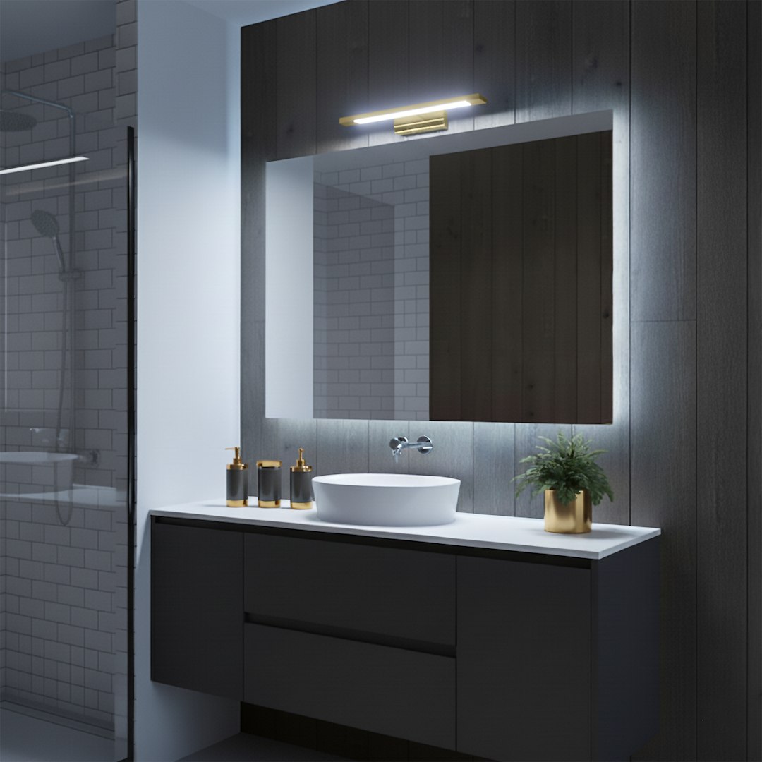 Kinkiet Klimt L40 LED 8W CCT IP44 w kolorze złotym, zamontowany nad lustrem. Oświetlenie ma prosty, nowoczesny design z metalowym wykończeniem, emitującym ciepłe światło. Widoczna aranżacja łazienki z umywalką i rośliną w doniczce.