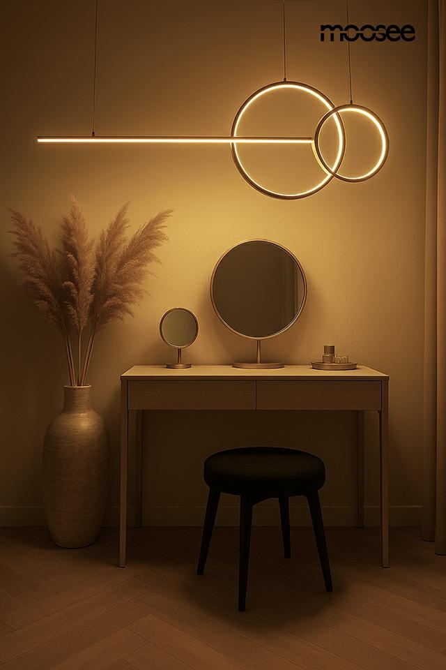 Lampa wisząca LED Rage 30W 3000K w złotym kolorze, zawieszona nad toaletką. Składa się z prostokątnego i okrągłego elementu. W tle widoczna stylowa toaletka, lustro oraz dekoracyjna roślina w wazonie. Ciepłe, miękkie światło.